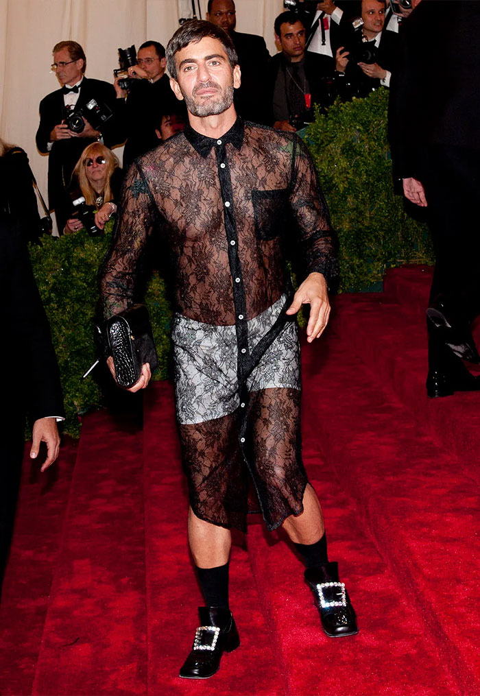 Male celebrity wearing a sheer lace outfit and holding a clutch, showcasing bizarre Met Gala outfits on the red carpet.