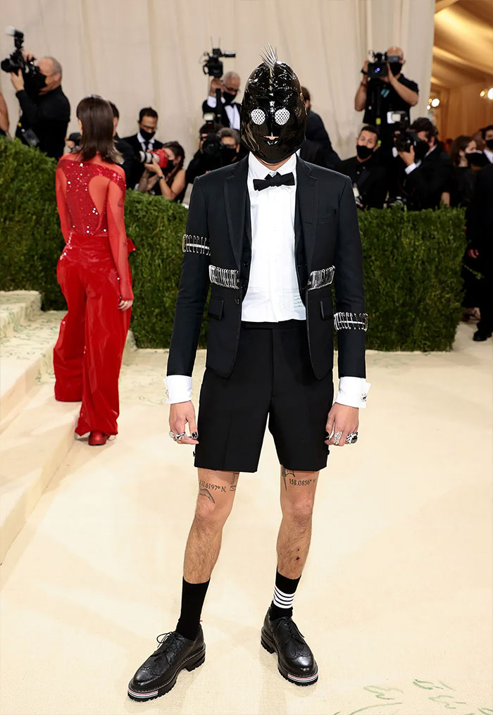 Celebrity in a unique mask and black suit with shorts posing amid photographers at an event with Met Gala fashion fails theme.