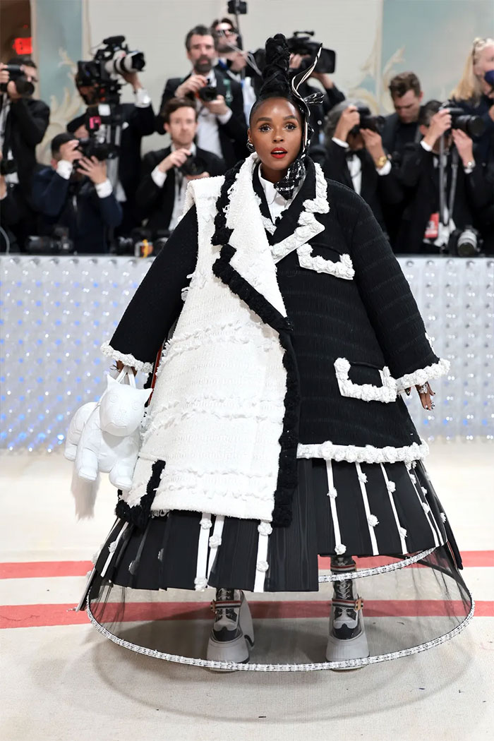 Celebrity in a bizarre black and white Met Gala outfit with avant-garde design and photographers in the background.