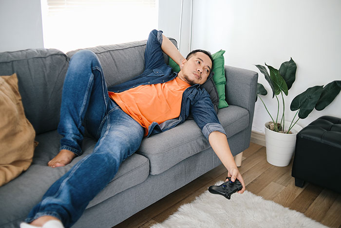Roommate's boyfriend relaxing on a gray sofa with a gaming controller in hand, in a modern living room.
