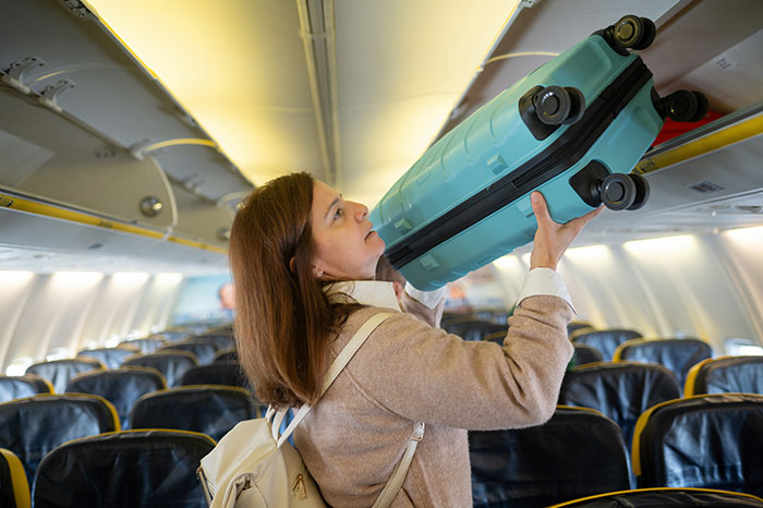 Woman placing luggage in overhead compartment on a plane, illustrating perfect revenge travel scenario.