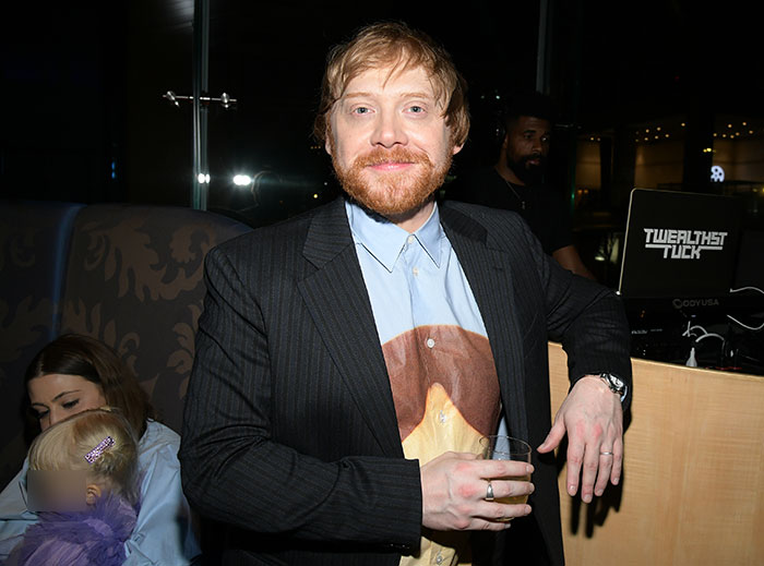 Rupert Grint, smiling at an event, with family in the background.