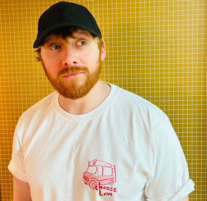 Actor in a cap and "Choose Love" shirt against a yellow tiled background.