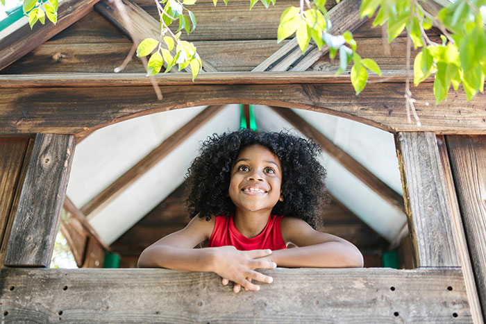 Young girl smiling inside a childhood tree house surrounded by green leaves and wooden structure outdoors.
