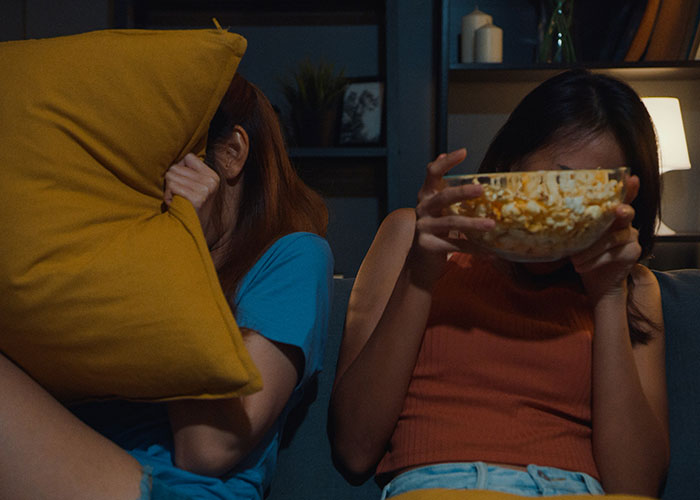 Two people on a couch, one hiding behind a pillow, the other holding popcorn, reacting to a scene.