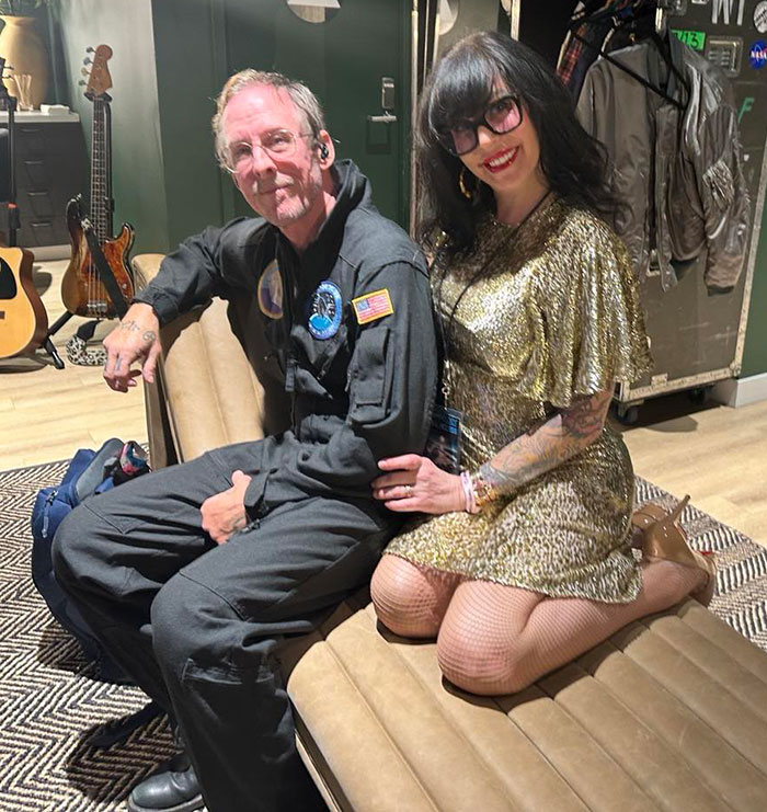 Man and woman sitting on a couch in a music studio, with guitars visible in the background.