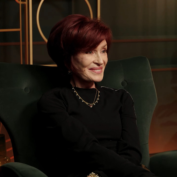A woman seated on a green chair, wearing a black outfit and necklace, with an elegant hairstyle and a smile.