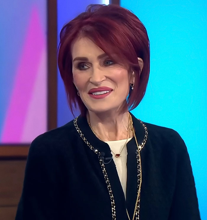 A woman with red hair in a black jacket speaks on a blue background, related to Ozempic feet side effect.