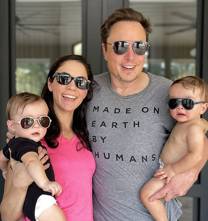A smiling family with man wearing sunglasses and a "Made on Earth by Humans" shirt, holding babies in sunglasses.