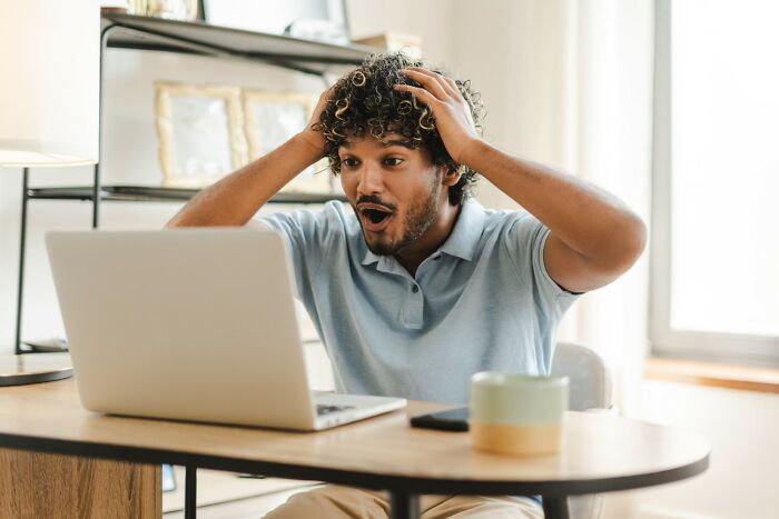 Shocked man at a desk reacting to traitor discussions online, hands on head, in a casual room setting.