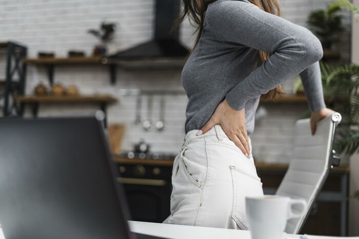 Woman enduring unexplainable pain, holding her lower back while standing in a modern kitchen with a laptop nearby.