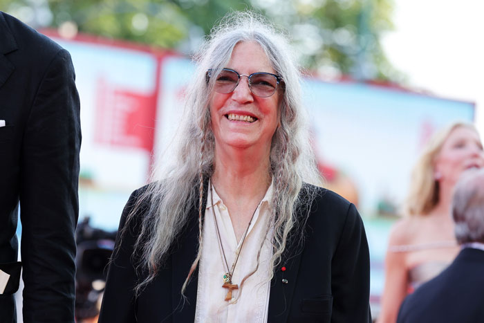 Punk rock legend smiling outdoors, wearing glasses and a black jacket.