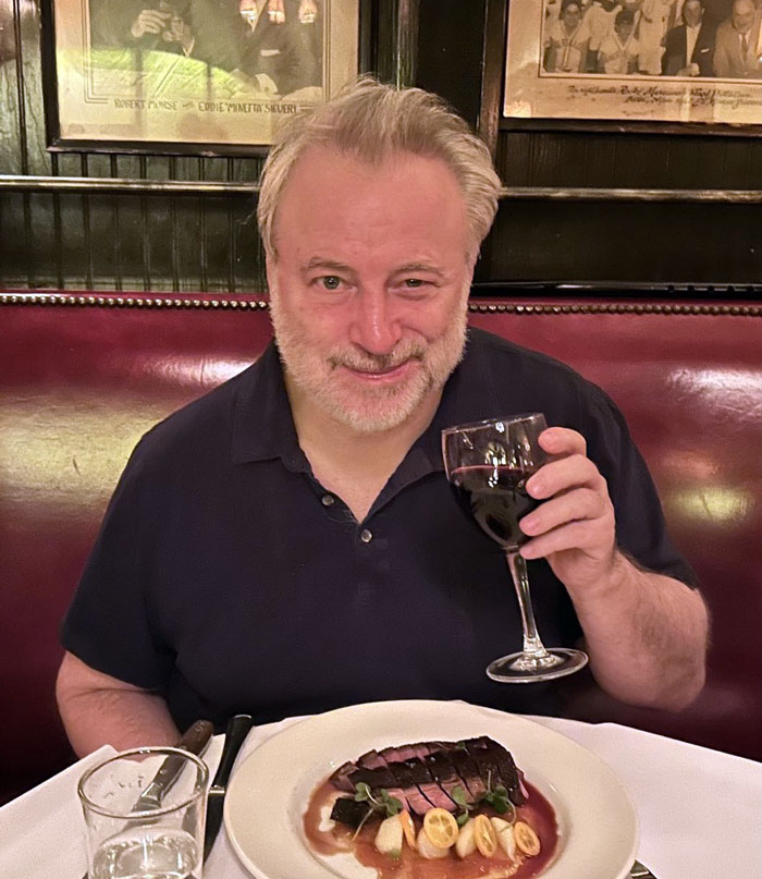 Man in black shirt holding a wine glass at a restaurant, with a dish in front of him, referencing punk rock legend Patti Smith.