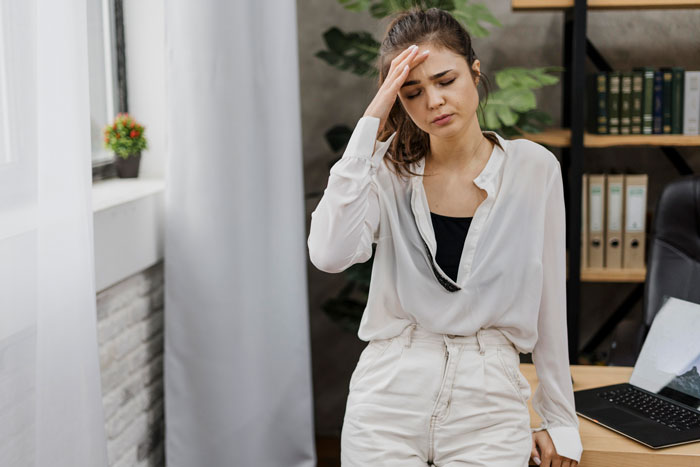 A woman in distress, wearing white, holds her forehead in an office setting, illustrating personal relationship struggles. A woman in distress, wearing white, holds her forehead in an office setting, illustrating personal relationship struggles.
