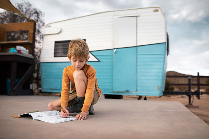 Child drawing outdoors near a blue and white trailer, focusing intently.