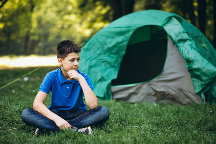 Boy in a blue shirt sitting on grass beside a green tent, outdoors in a park setting.