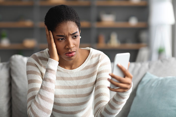 Woman in a striped sweater looks frustrated while reading a message on her phone related to babysitter stress.
