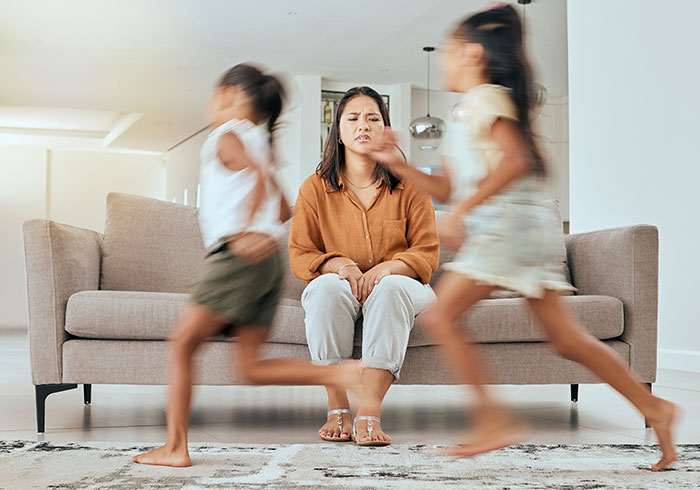 Woman sitting stressed on a couch while two kids run around, reflecting stress in a babysitting scenario.