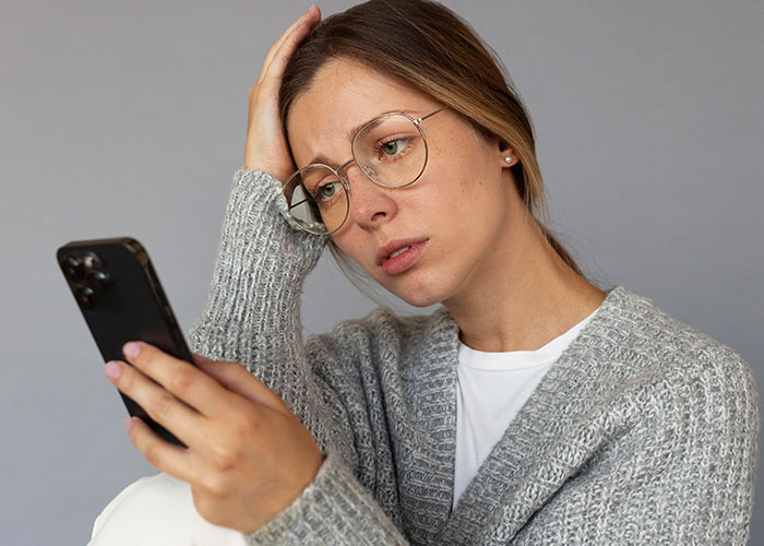 A worried woman with glasses looking at her phone, regretting family drama and $2,000 damages.