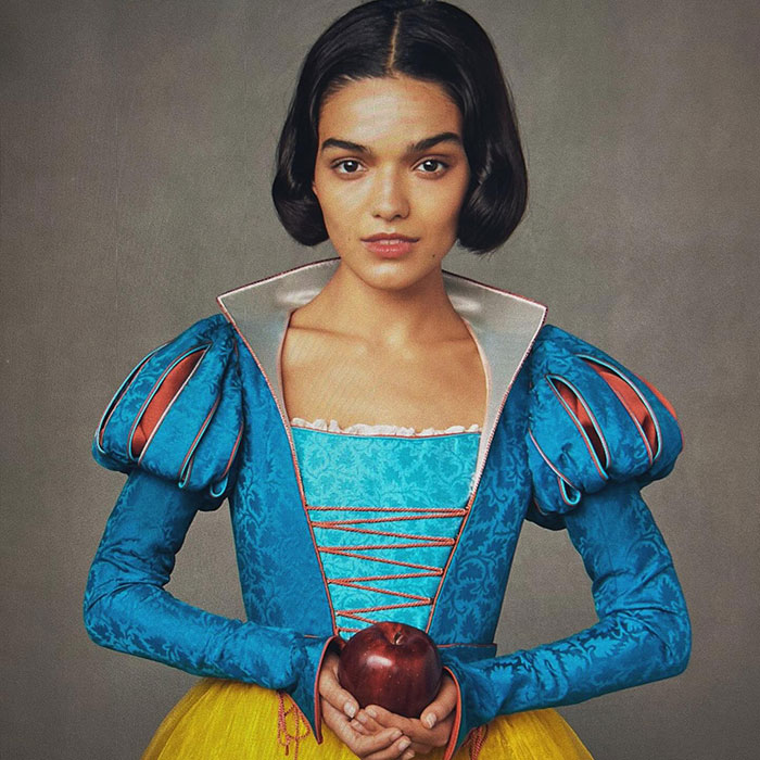 Young woman in a vibrant costume, holding an apple, spotlight on "Rachel Zegler.