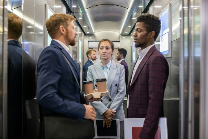 Three people in a business elevator, holding coffees and chatting, conveying legal but psychopath vibes.