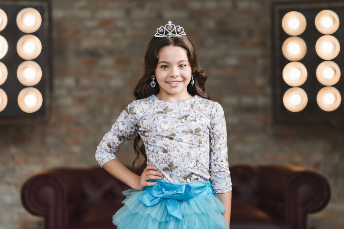Young girl in a floral top and blue tutu, wearing a tiara, standing confidently in a studio setting.
