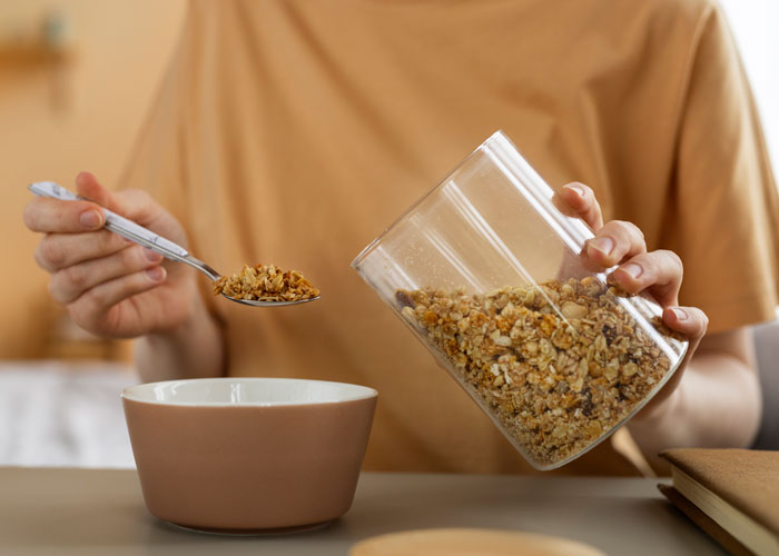 Person pouring cereal into a bowl with a spoon, in a kitchen setting, illustrating a daily routine.