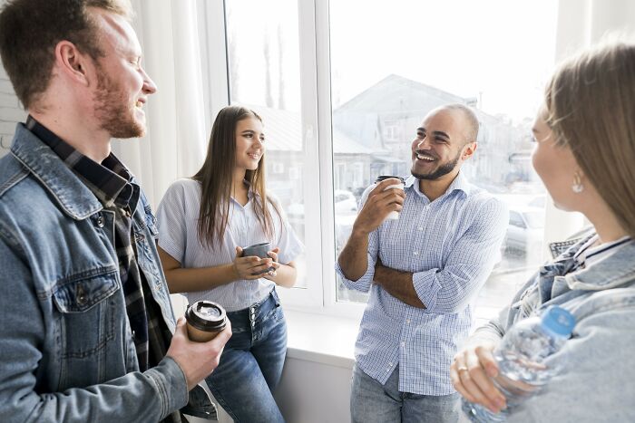A group of people casually chatting and smiling by a window, each holding drinks, in a relaxed indoor setting.
