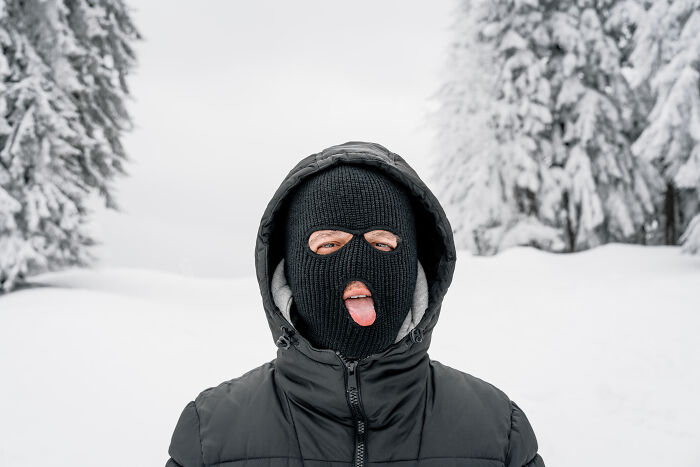 Person in a ski mask sticks out tongue in snowy forest, conveying psychopath vibes despite legality.