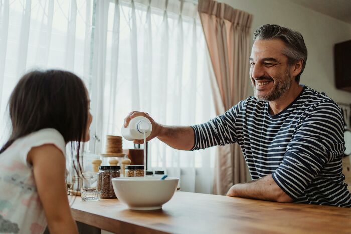 Man pouring milk while smiling at a girl at the table, evoking legal yet unsettling vibes.
