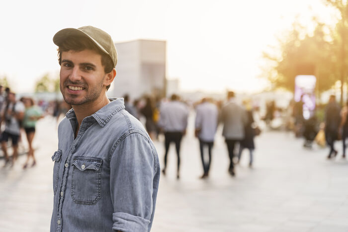 Young man in a cap and denim shirt smiles outdoors, with people walking in the background.