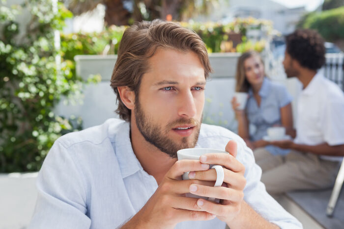 Man sipping coffee outdoors, looking thoughtful, with two people chatting in the background, conveying subtle psychopath vibes.