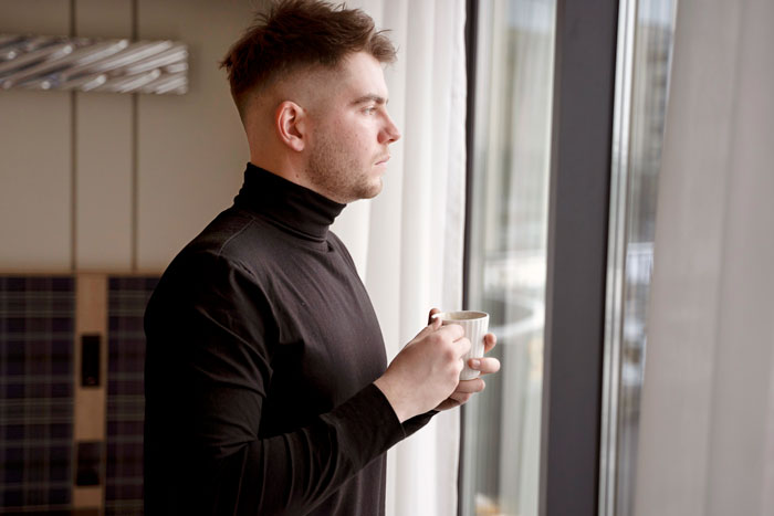 Teen boy in black turtleneck holding cup, looking out window thoughtfully, capturing moment related to dad catching son creeping.