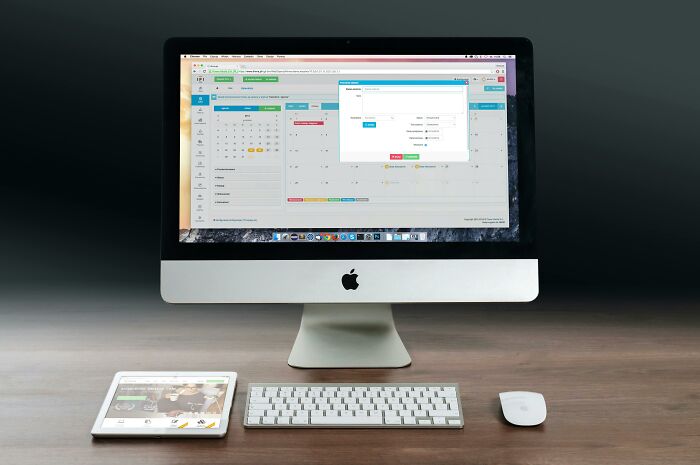 iMac on a desk showing a calendar app, with a keyboard, mouse, and a tablet displaying a website, explain-adult-moments.