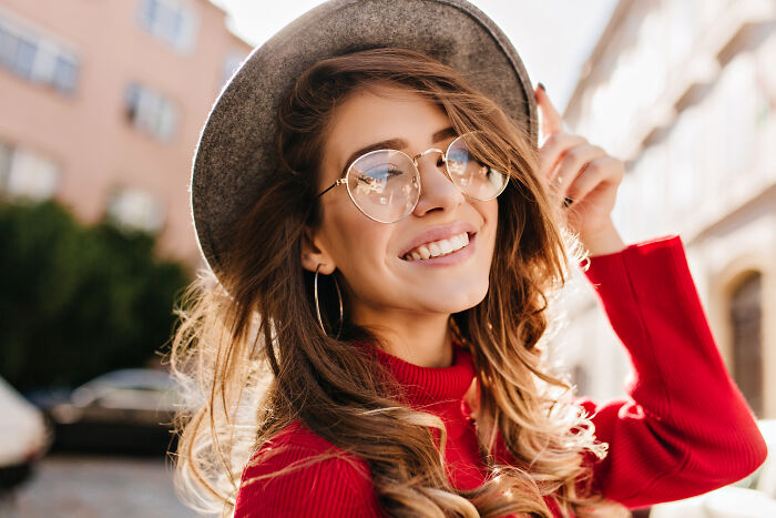 Smiling woman in glasses and a hat, embodying joyful adult moments in a city setting.