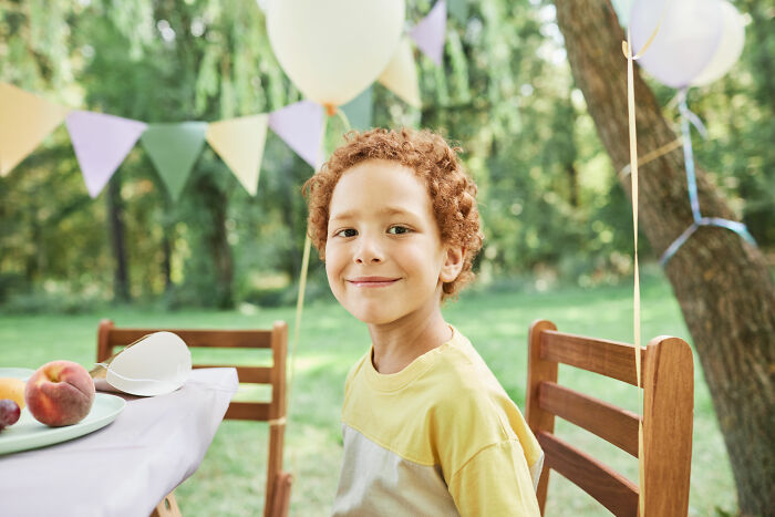 Child at birthday party in outdoor venue, smiling, with balloons and table setting in background.