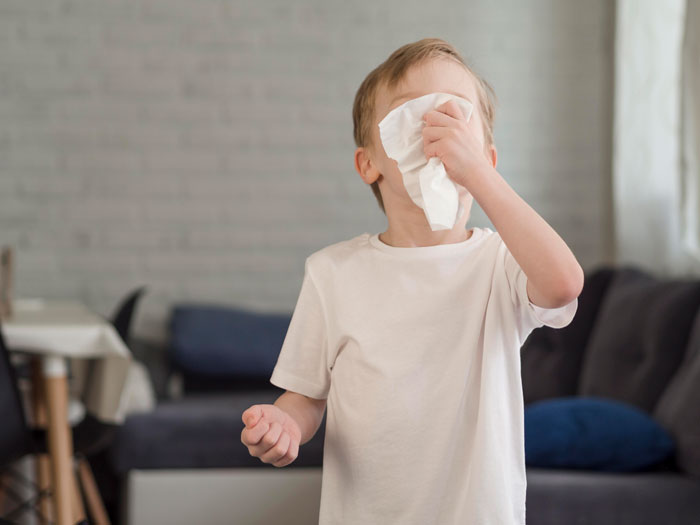 Young boy with allergies wipes face with tissue in living room.