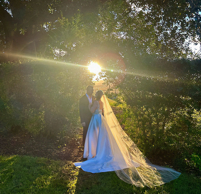 Burns survivor at wedding with partner, sunlight streaming through trees, creating a magical backdrop.
