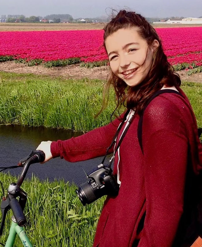 Former child star smiling on a bicycle, wearing a red sweater, with a camera around her neck in a flower field.