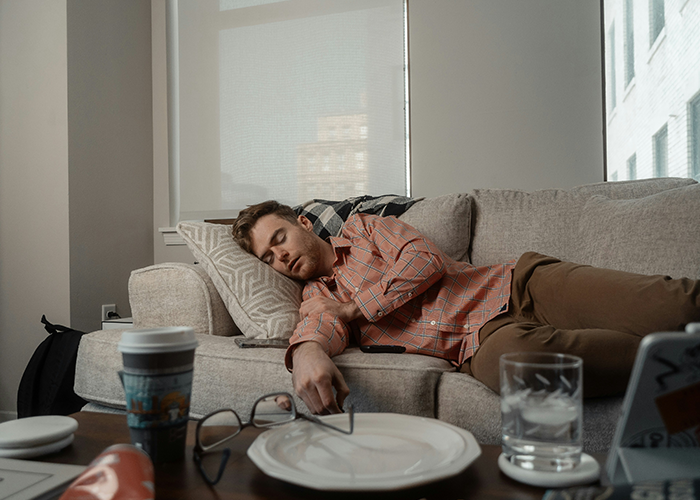 Man sleeping on couch amid household chores, with glasses, coffee, and plate nearby, representing chores and rent themes.