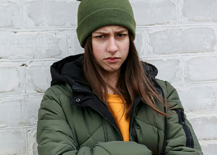 Young woman in a green jacket and beanie, looking upset in front of a brick wall, representing tension over chores and rent.