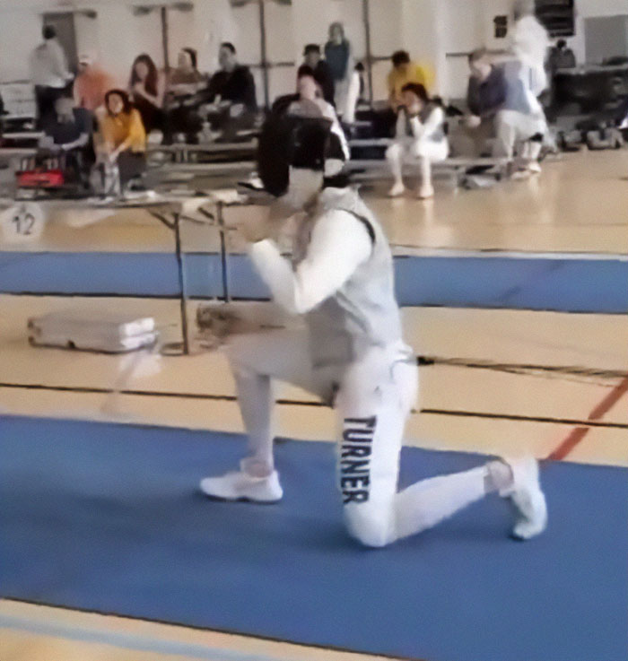 Trans athlete kneeling in fencing gear on a blue mat during a college fencing event.