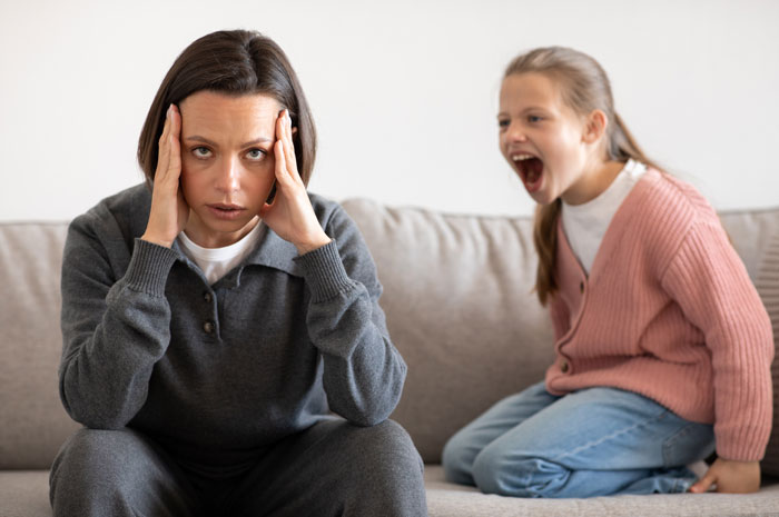 Stepmom stressed on sofa while child in pink cardigan shouts, depicting family tension.