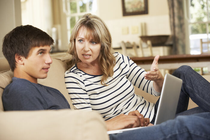 Stepmom having a serious discussion with teenage stepson on the sofa, holding a laptop, in a well-lit living room.