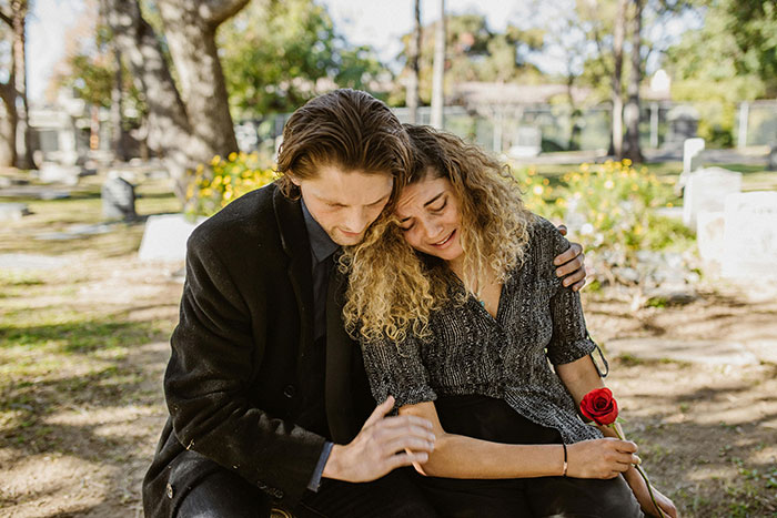 A couple seated in a cemetery, sharing a comforting embrace, symbolizing emotional karma amidst past family turmoil.