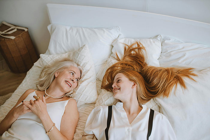 Stepmom and stepdaughter lying on a bed, smiling at each other, surrounded by white pillows.