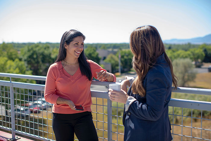 Two women casually talking on a balcony, one in a peach top holding a phone, with a green landscape in the background.