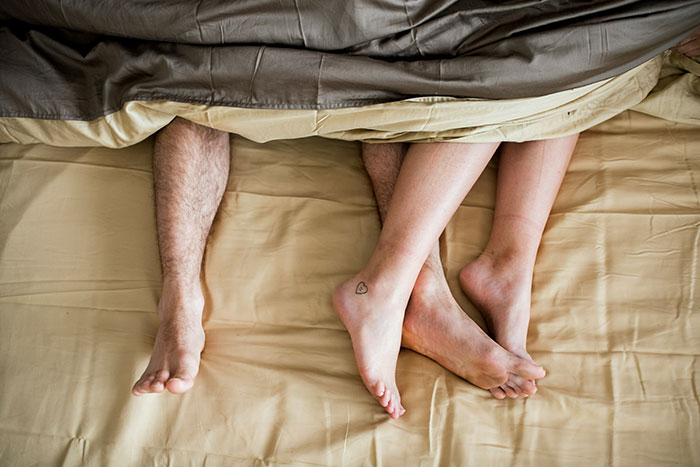 Two pairs of legs intertwined under a blanket, one with a heart tattoo, signifying a crazy casual admission scenario.