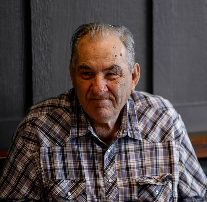 Elderly man in plaid shirt smiling casually, evoking tales of crazy admissions and life stories.