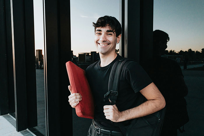 Smiling person holding a red item, wearing a black shirt and backpack, casually in front of a building.